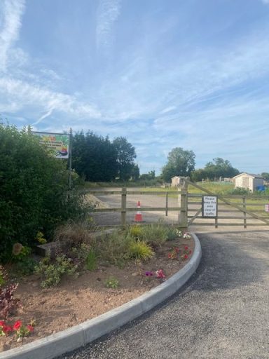 Entrance the Wolston Allotments Entrance to a gravel path surrounded by greenery and flowerbeds under a clear blue sky.