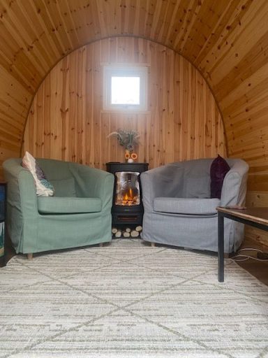 Inside the Therapeutic Camper Cosy interior with two armchairs, a small table, and a wood stove against a wooden wall.