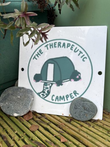 Logo of The Therapeutic Camper. Logo for "The Therapeutic Camper" featuring a tent and rocks, set against greenery.