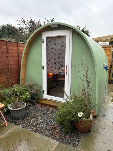 The Therapeutic Camper Pod Green pod-shaped garden shed with a door and plants around it.