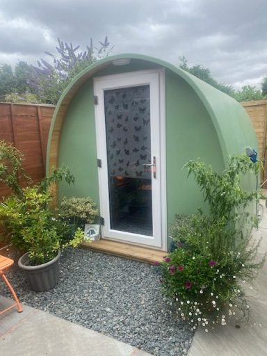 The Therapeutic Camper Pod Small green pod-shaped cabin surrounded by plants and gravel, with a wooden entrance.